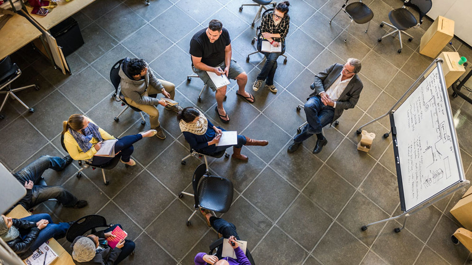 Students gather in a circle around a professor and jot down notes during the Design Methods course.