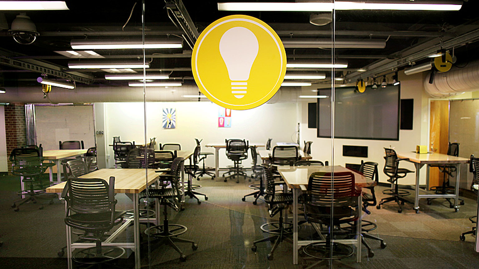 A view of a classroom full of adaptable rolling furniture and a large icon of a yellow lightbulb on the glass wall in center-screen.